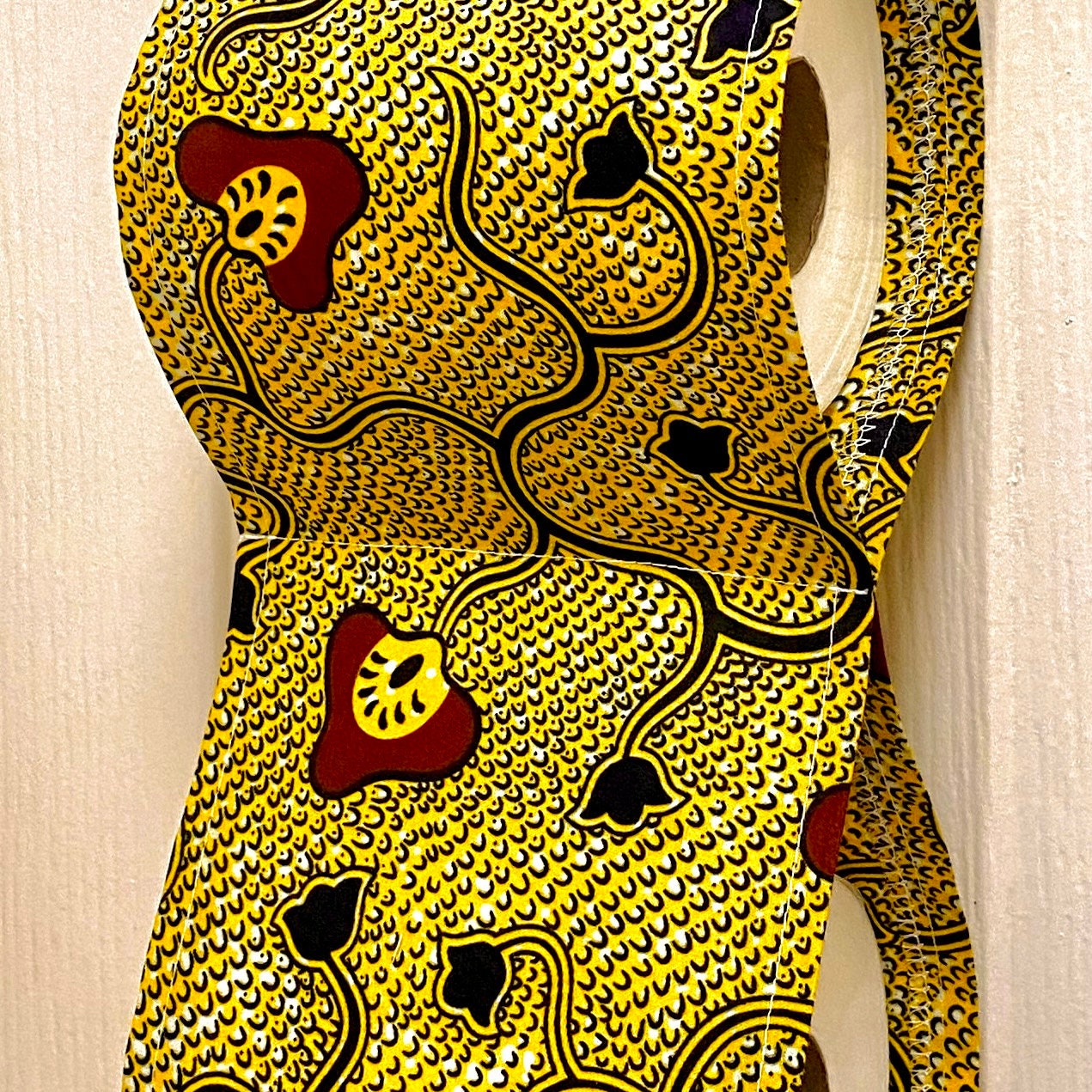 Close up of African wax print cotton toilet roll holder in yellow and black with mushroom and flower motifs, hanging in a bathroom.
