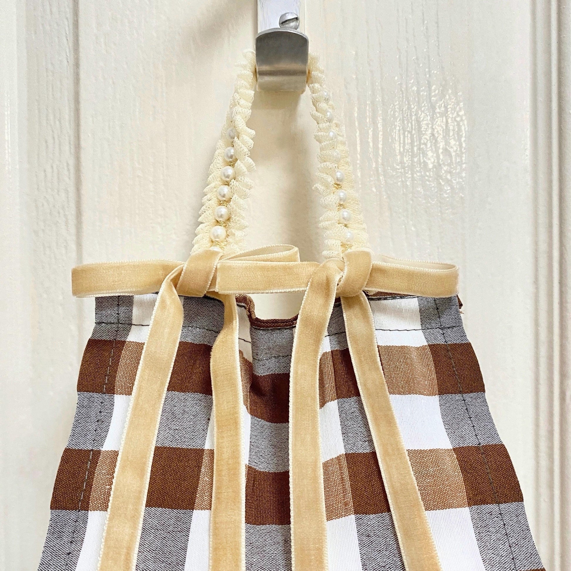 Close up brown and white gingham fabric toilet roll holder with velvet bows hanging in a bathroom setting.