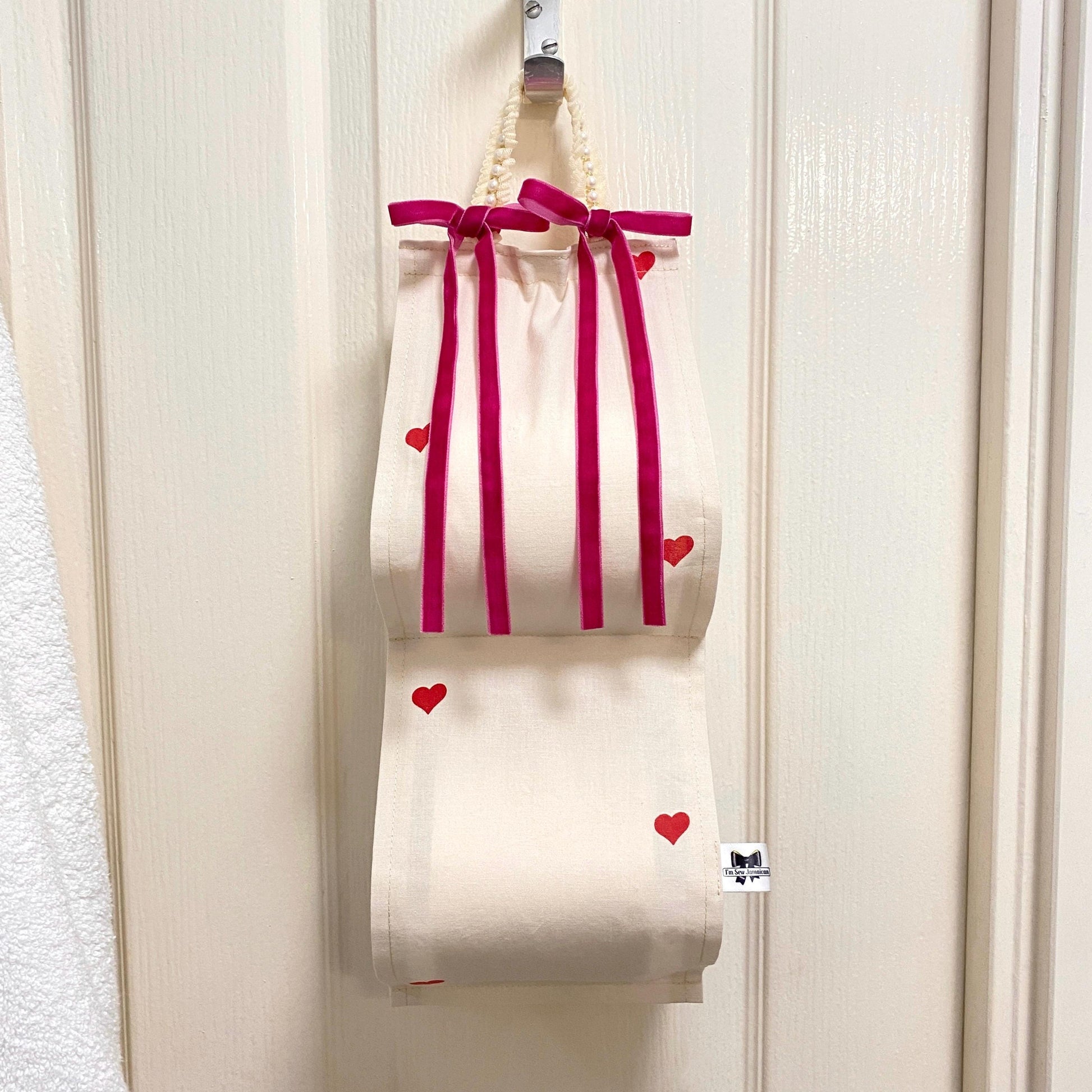 Cotton fabric toilet roll holder with small hearts and pink bows hanging behind the bathroom door, front view.