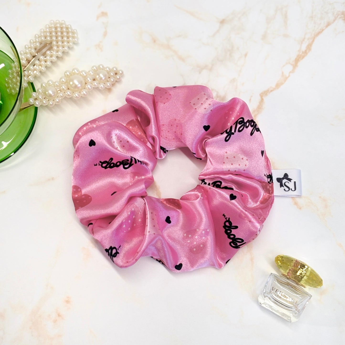 Pink Betty Boop satin scrunchie in a flat lay with props, including hair accessories and perfume.