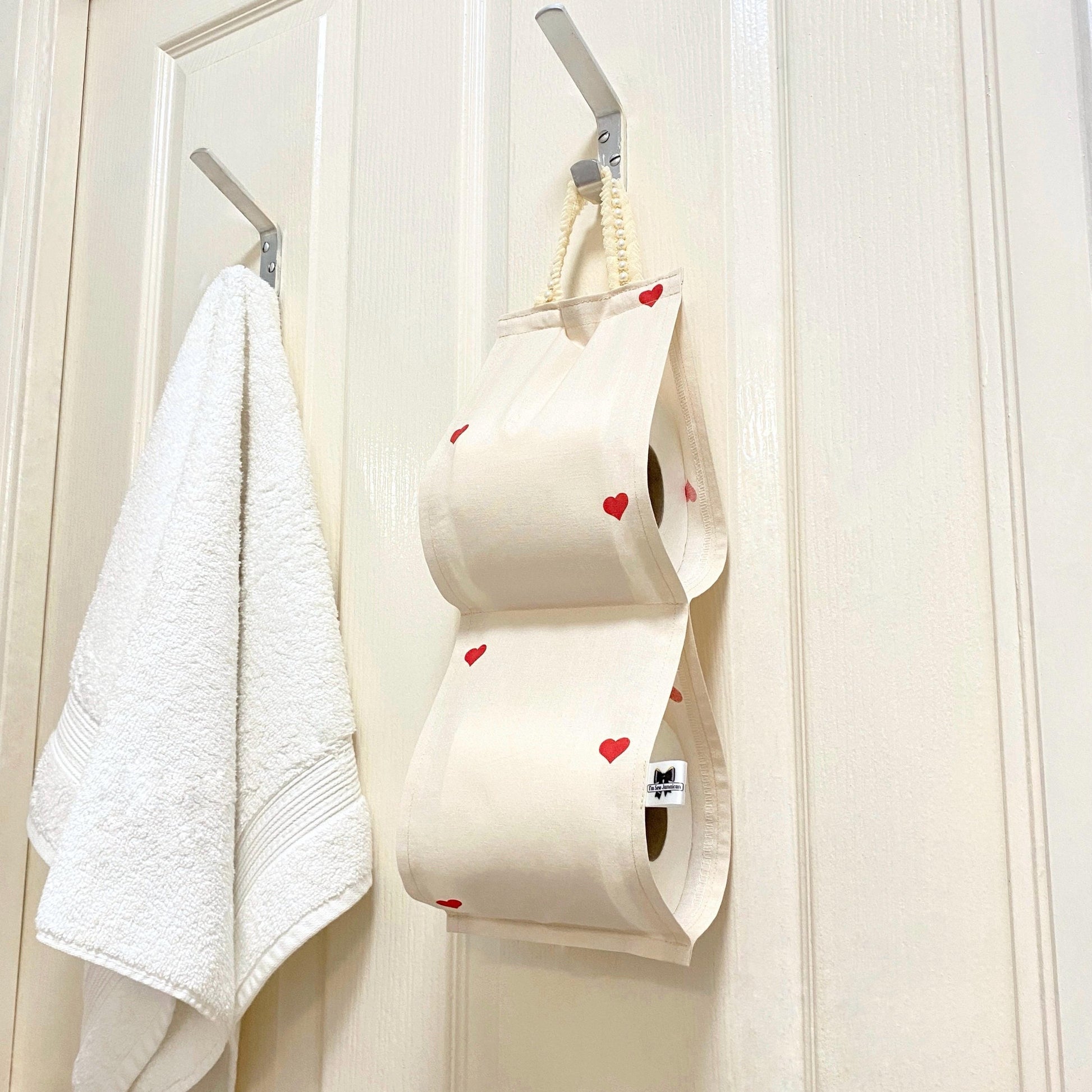 Cotton fabric toilet roll holder with hearts hanging behind the bathroom door beside white hand towel.