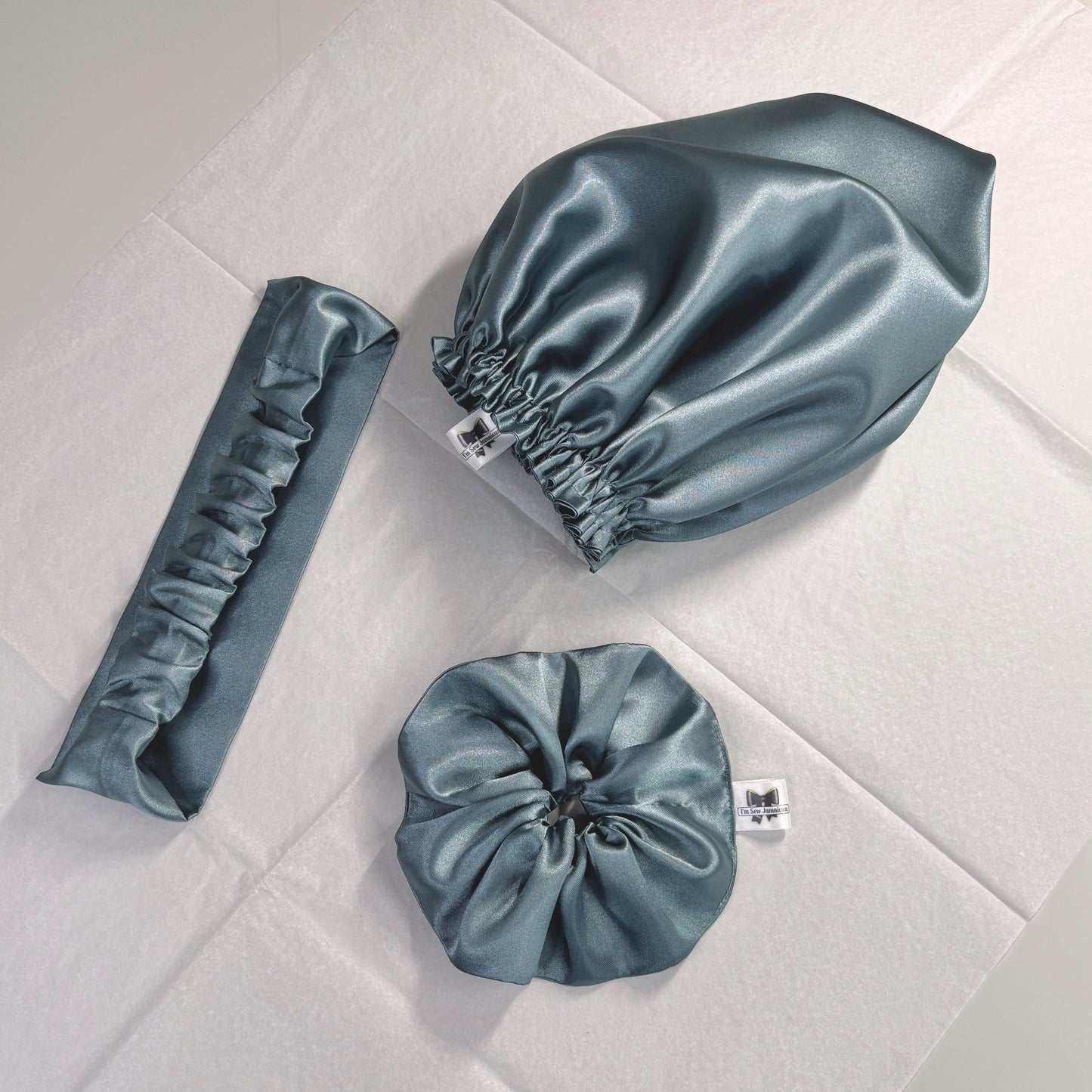 Aqua blue/green satin hair bonnet, satin headband, and satin hair scrunchie displayed on white tissue paper for customer.