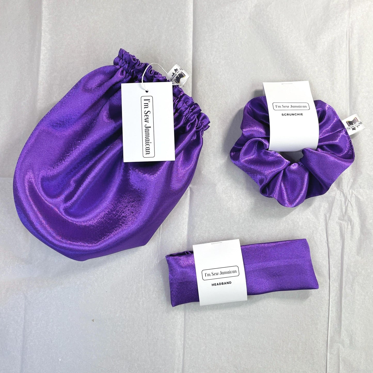 Purple satin hair bonnet, satin headband,  satin hair scrunchie displayed on brilliant white tissue paper with packaging.