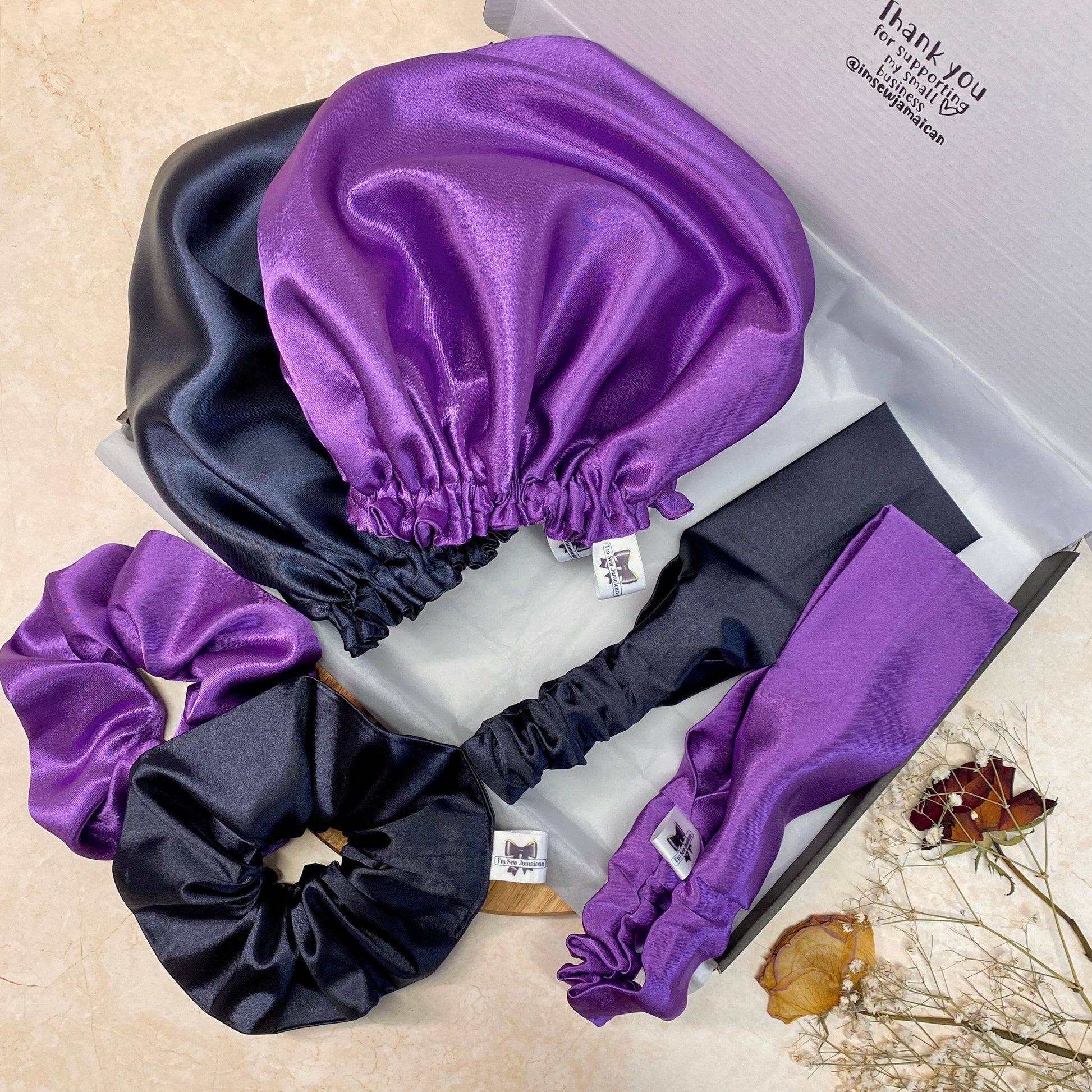 Purple and navy blue satin hair bonnet, satin headband, and satin scrunchie in  in a postal box with white tissue paper displayed on marble background.