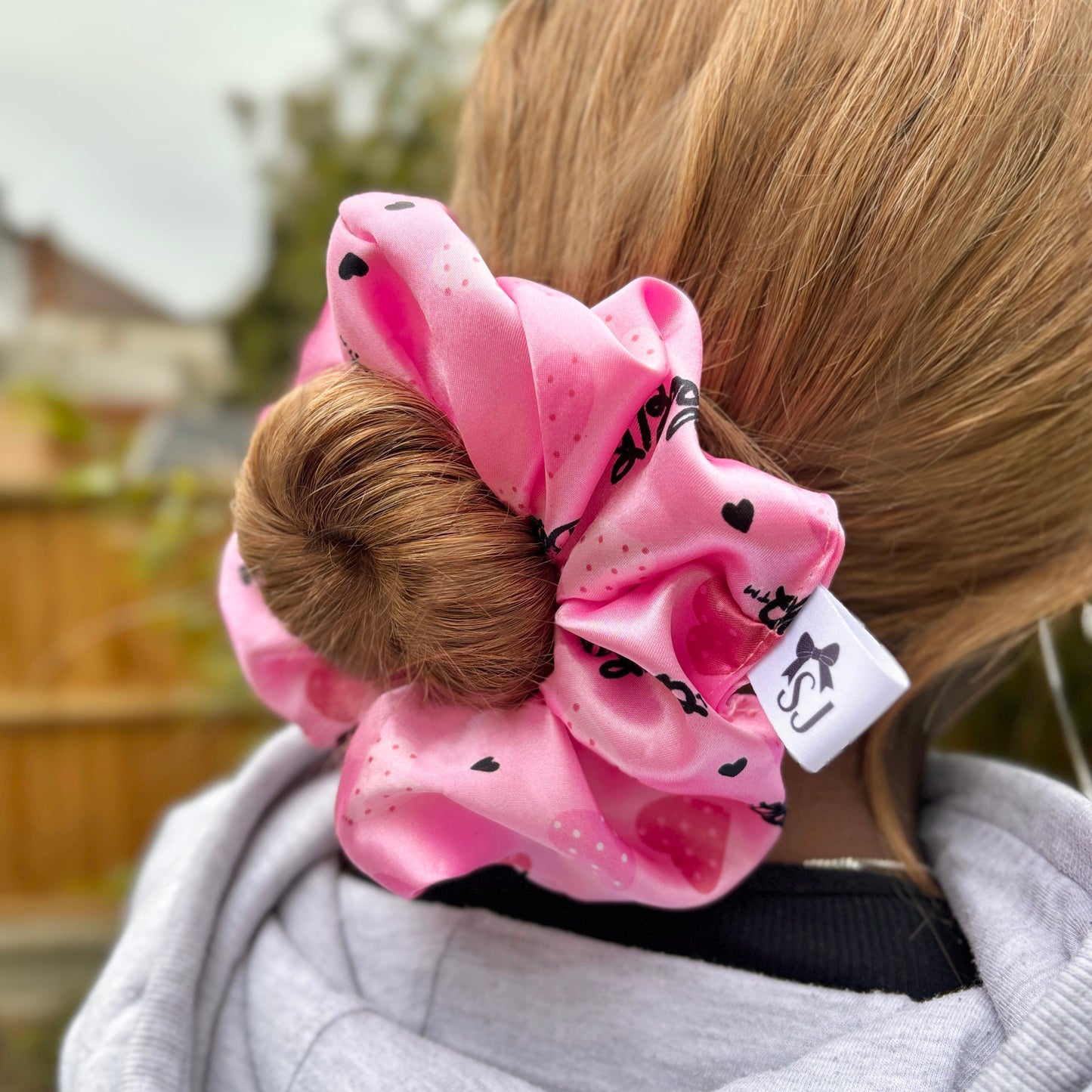 Side view pink Betty Boop satin scrunchie in a woman's blonde hair from the back, in front of a bamboo tree outside.
