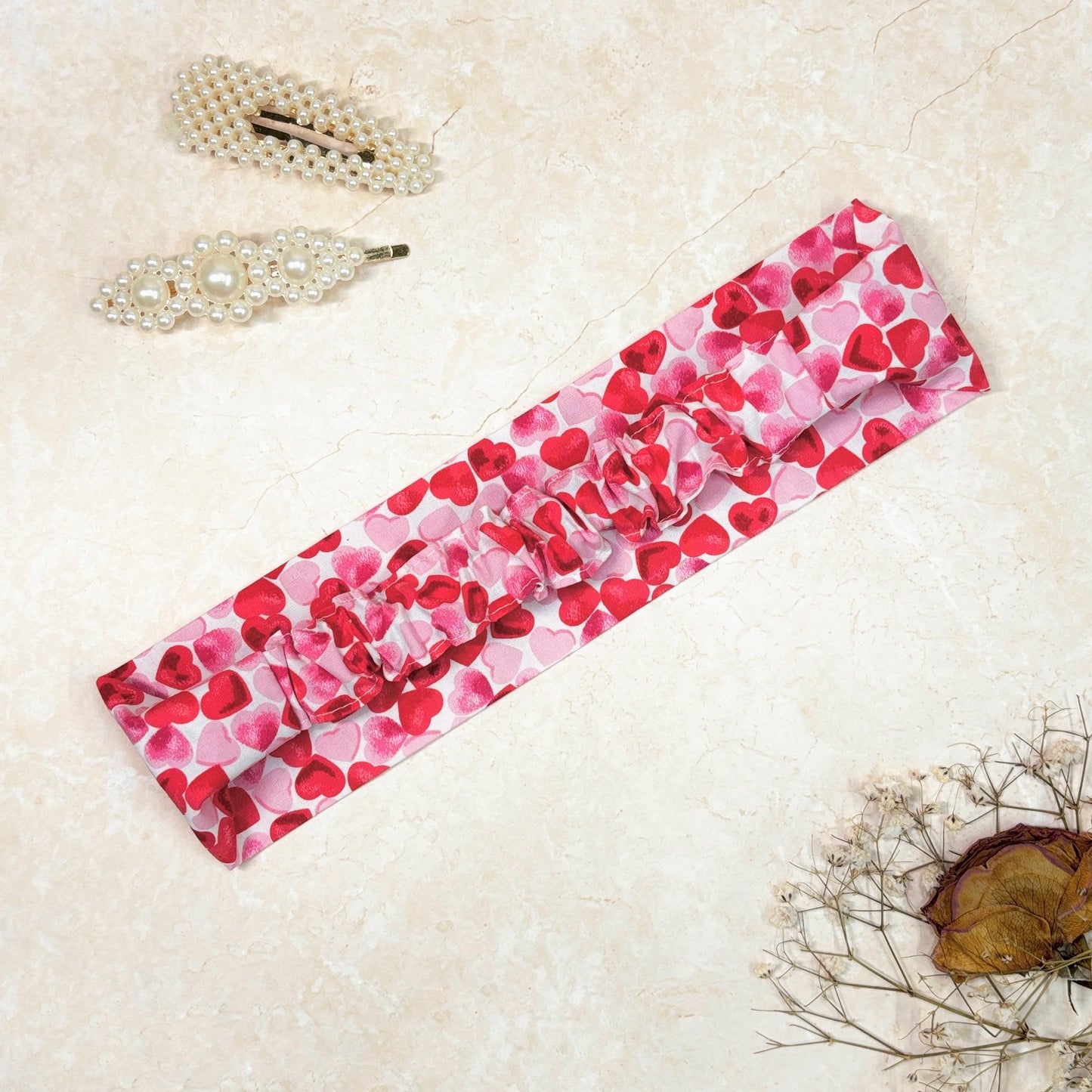 Pink and red heart print cotton headband, thick, flat, wide, side down with elastic ruffle in the back on a peach background with props, perfect for hair styling, fashion accessories, 