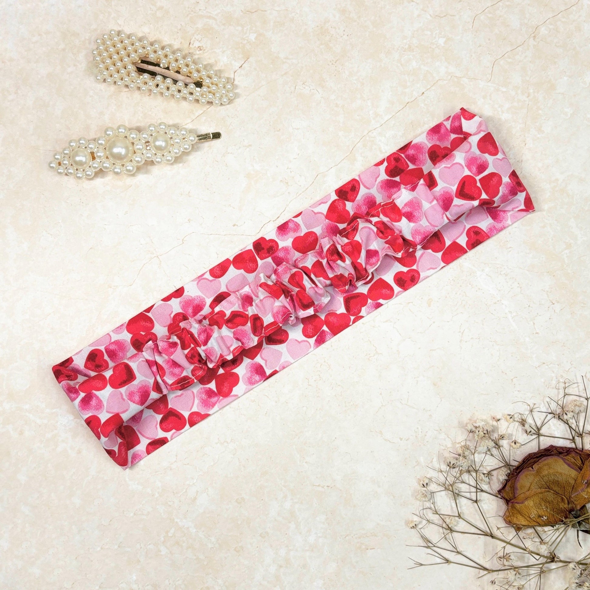 Pink and red heart print cotton headband, thick, flat, wide, side down with elastic ruffle in the back on a peach background with props, perfect for hair styling, fashion accessories, 