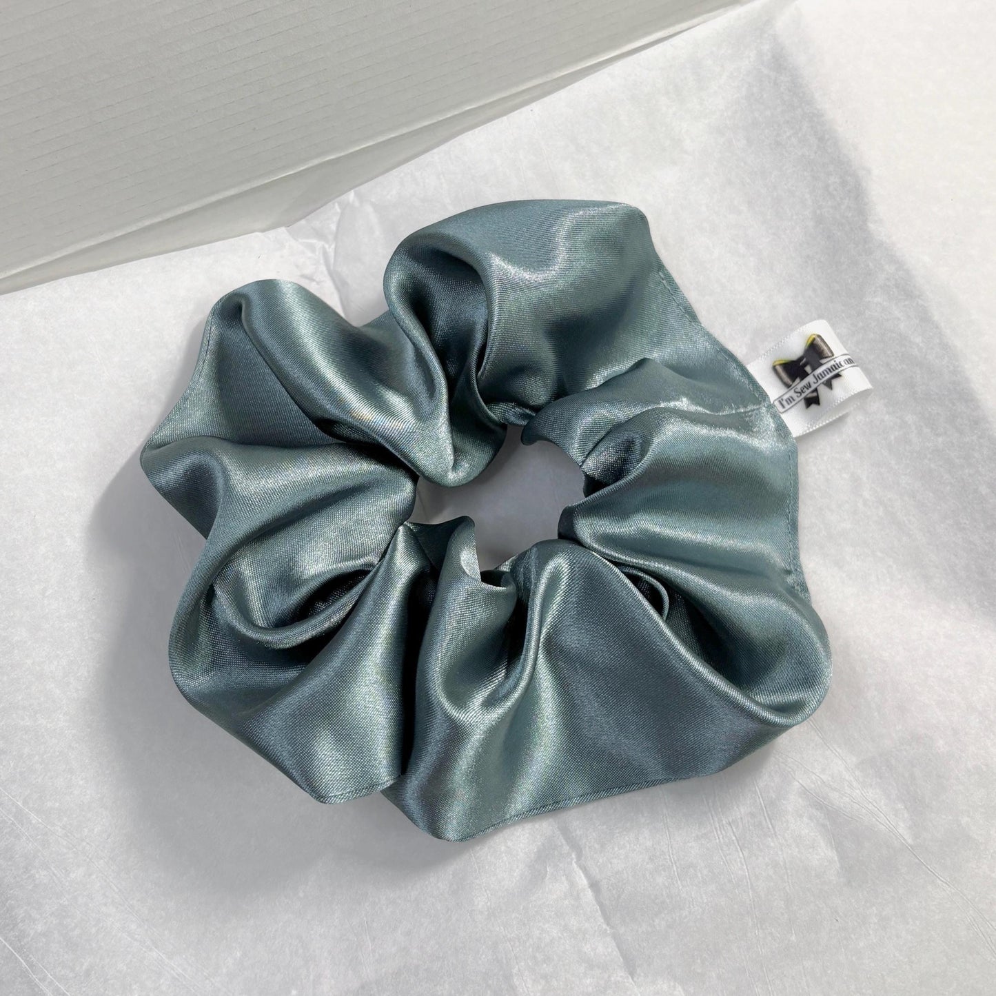 Aqua satin hair scrunchie with shine, stylish accessory for all hair types on white tissue paper on carboard box packaging.
