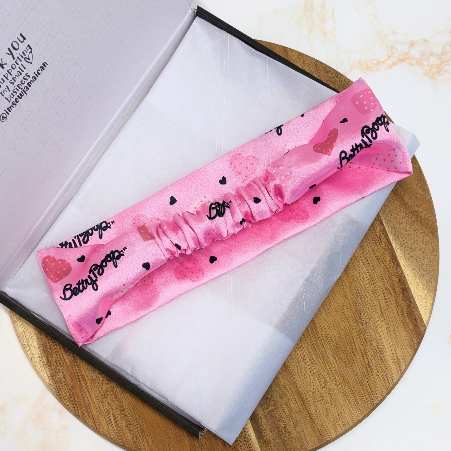 Pink Betty Boop thick wide flat headband facing downwards n branded packaging postal box on a dark brown wooden circle tray, perfect for hair styling, fashion accessories, and gifting from a small business.