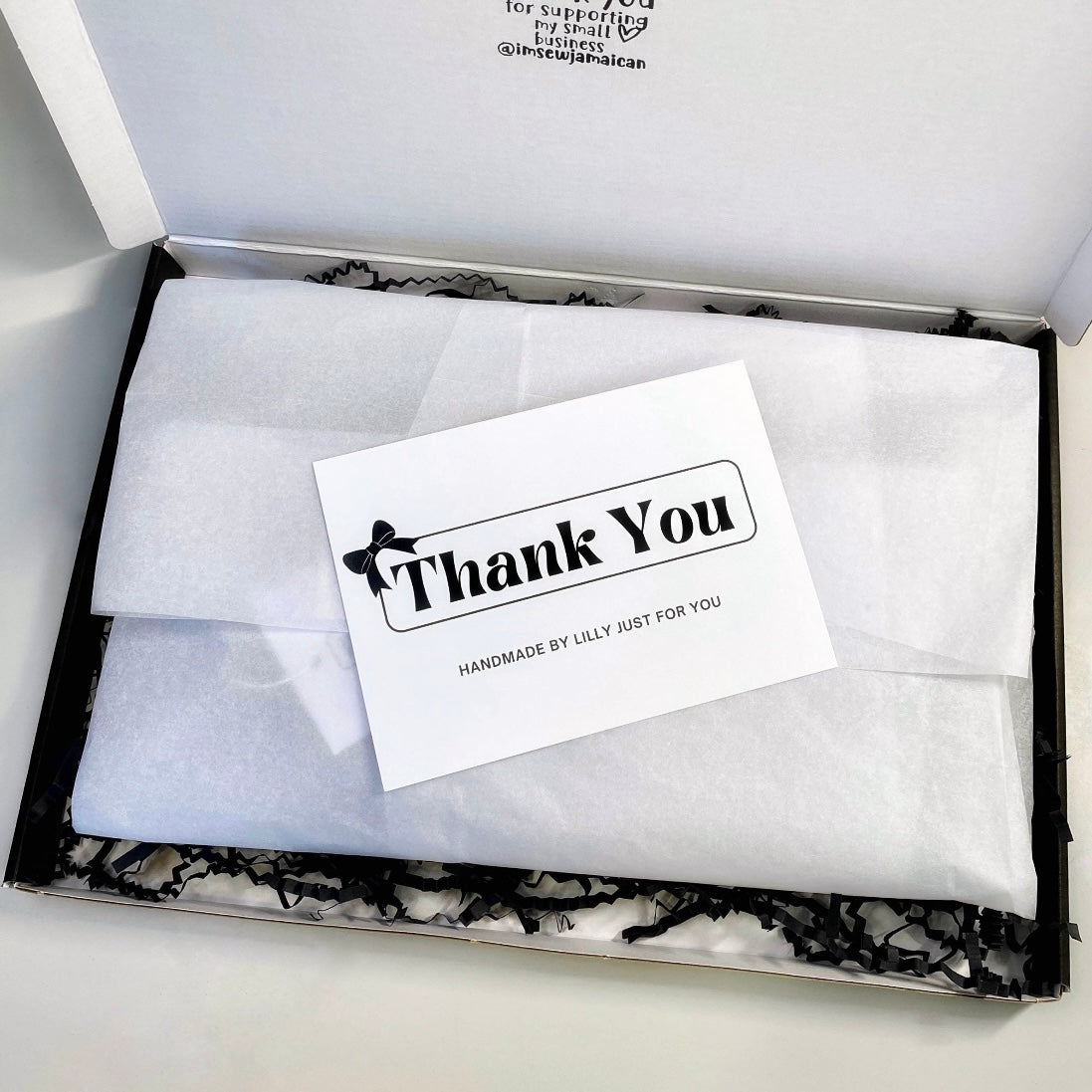 Small business gift packaging with white tissue wrap, black shredded paper, and a thank you card reading “Handmade by Lilly just for you” inside a branded postal box.