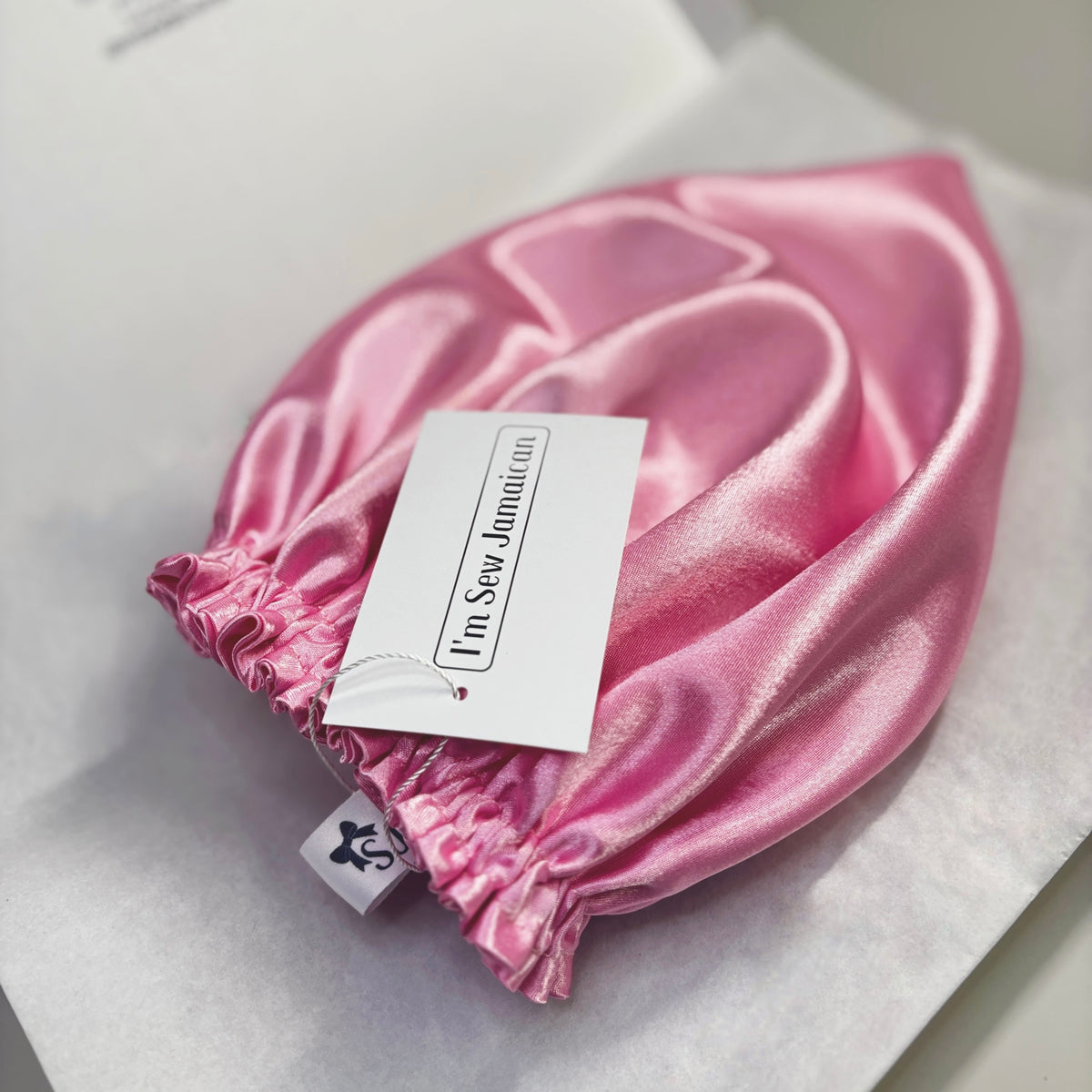 A shiny pink satin bonnet with an elasticized gathered edge, shown on white tissue paper with a paper hangtag that reads 'I'm Sew Jamaican'.