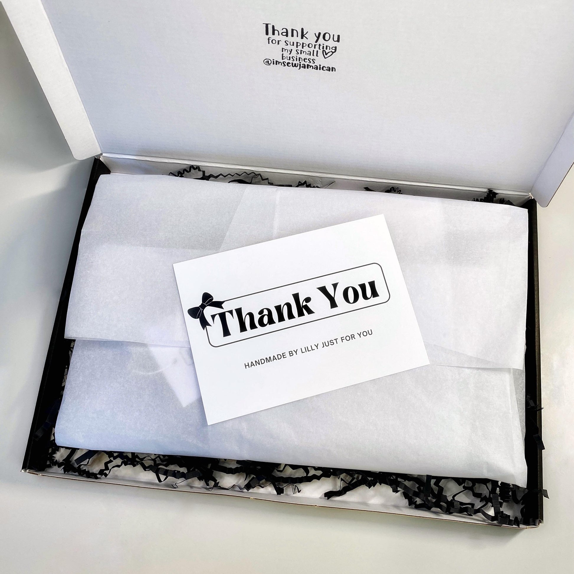 Small business gift packaging with white tissue wrap, black shredded paper, and a thank you card reading “Handmade by Lilly just for you” inside a branded postal box.