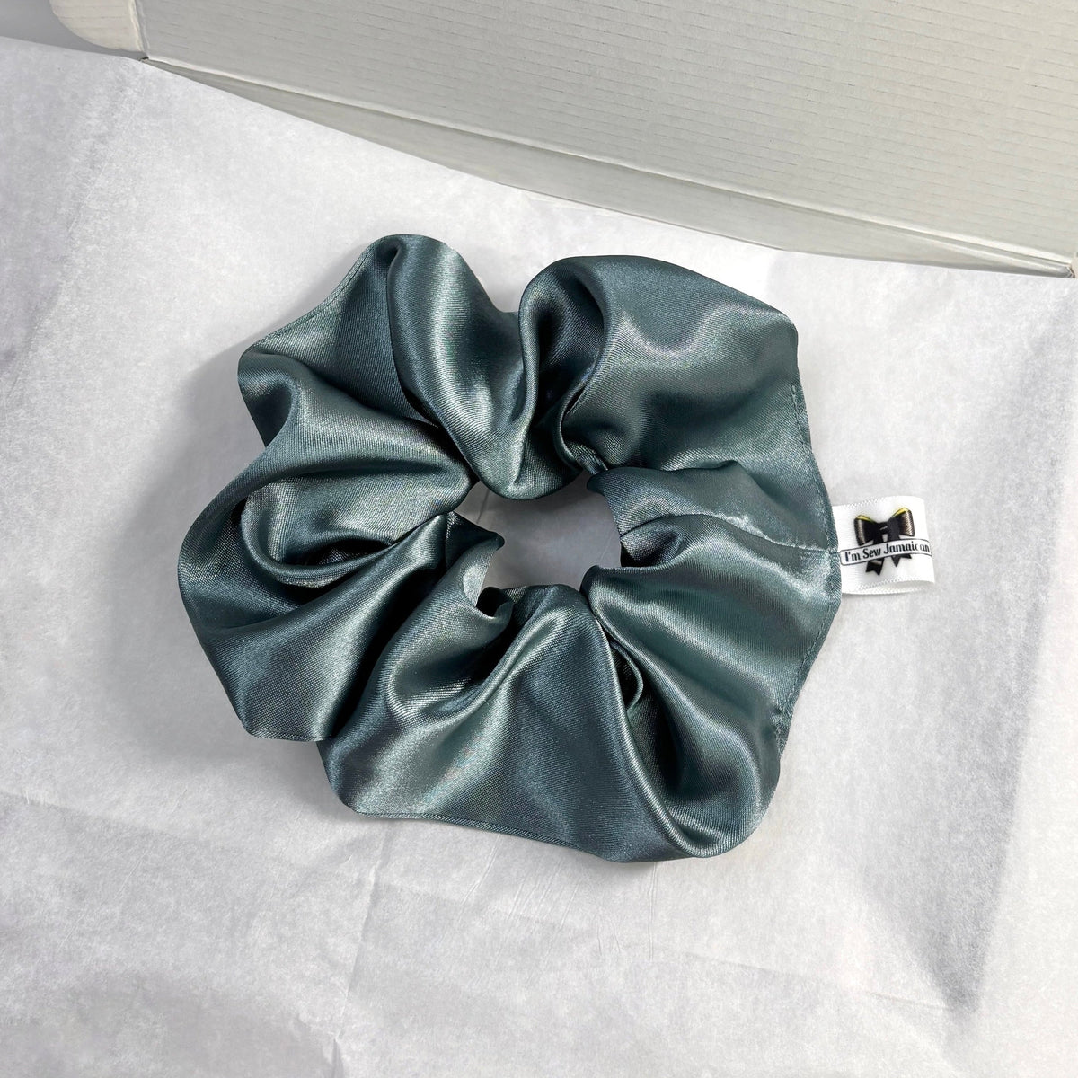 A dark teal satin hair scrunchie with a small white brand tag, displayed on white tissue paper next to its white gift box.