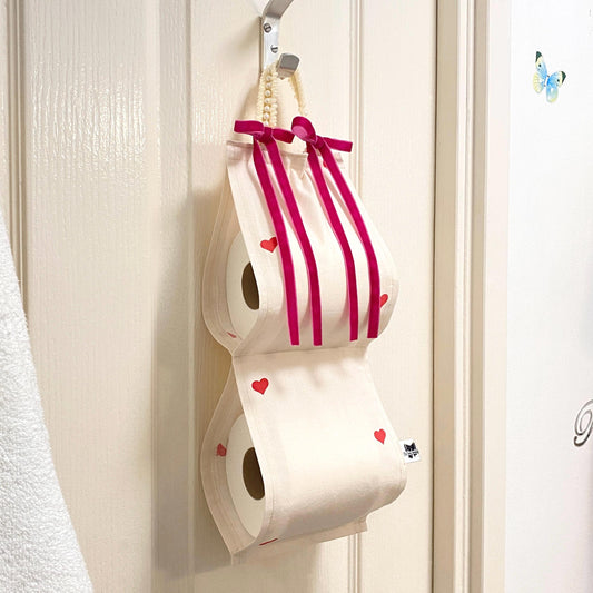 Cotton fabric toilet roll holder with hearts and pink bows hanging behind the bathroom door, side view.
