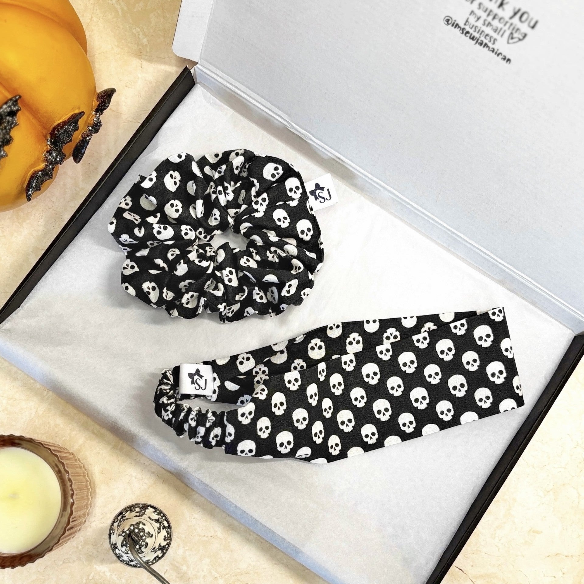 Black and white skull-patterned scrunchie and headband on a white box with a candle and pumpkin.