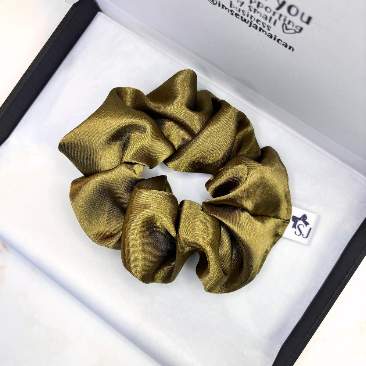 A shiny olive-gold satin scrunchie resting inside an open white jewelry box with a black border. A small white tag with the initials 'SJ' is attached to the scrunchie.