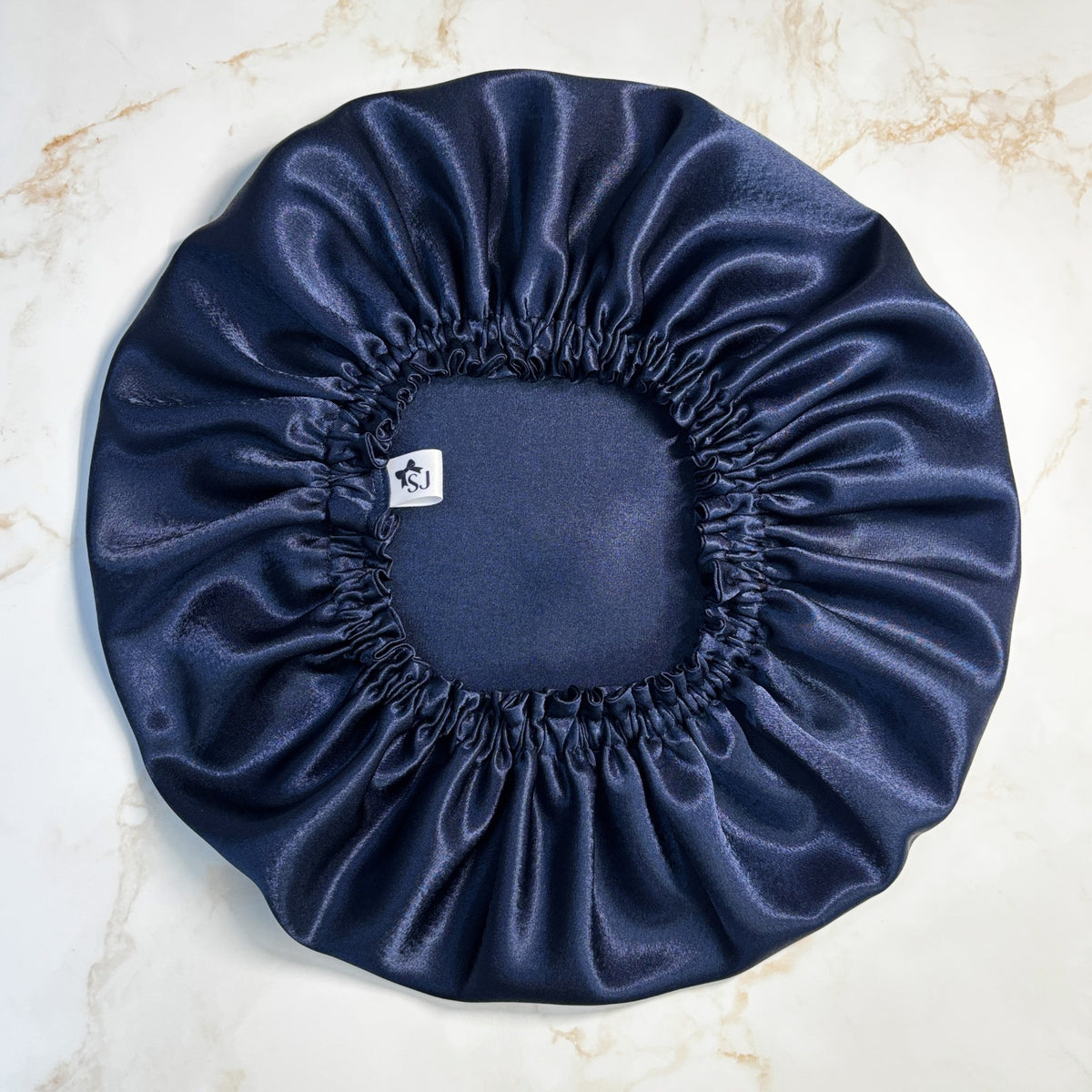A navy blue satin sleep cap or bonnet with an elasticized gathered edge, laid flat on a light beige marble surface. A small white brand tag is visible near the center.