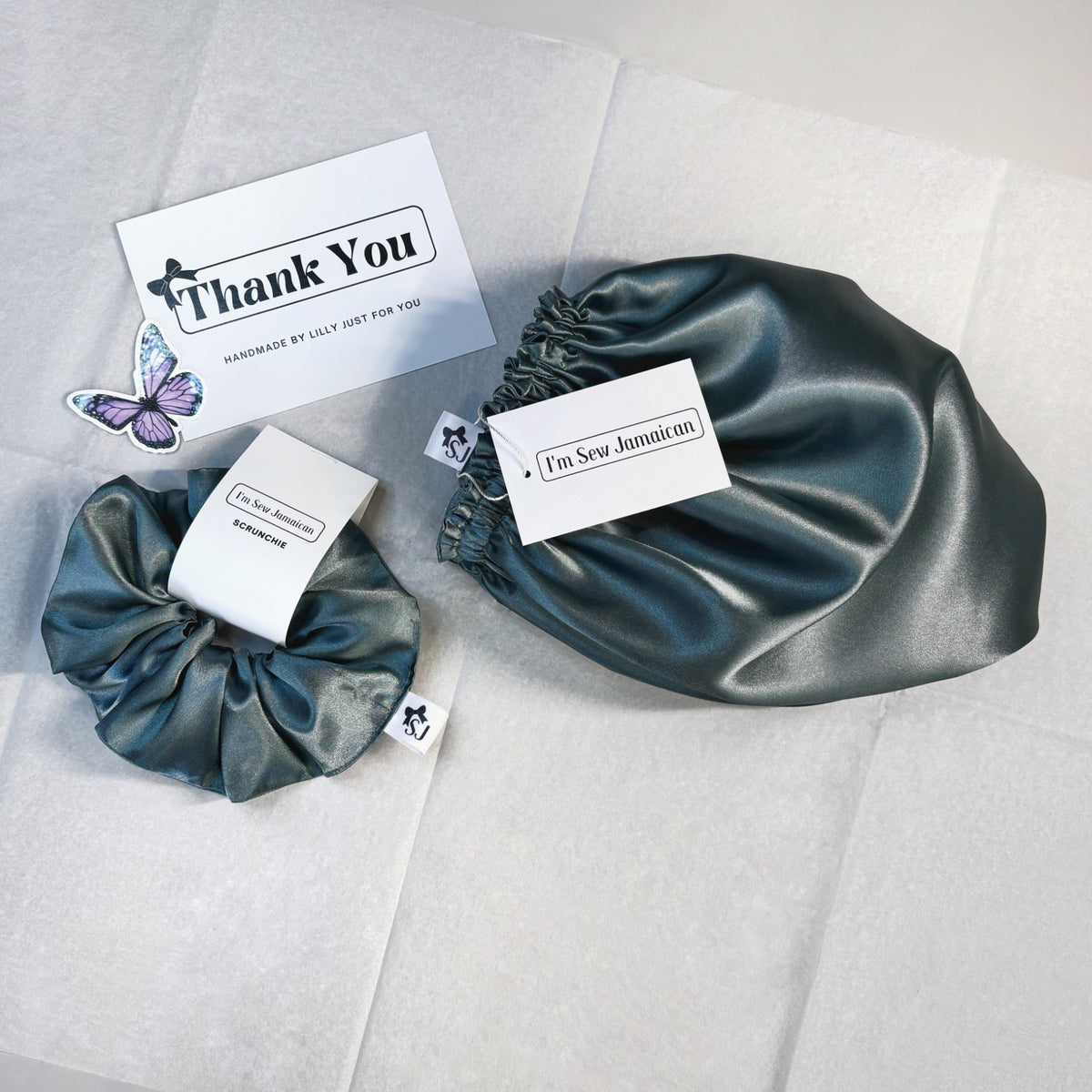 Two satin hair scrunchies in a dark teal-blue color are displayed on a white tissue paper background, each with a white paper tag. One tag reads 'I'm Sew Jamaican' and the other says 'Thank You' with a butterfly illustration.