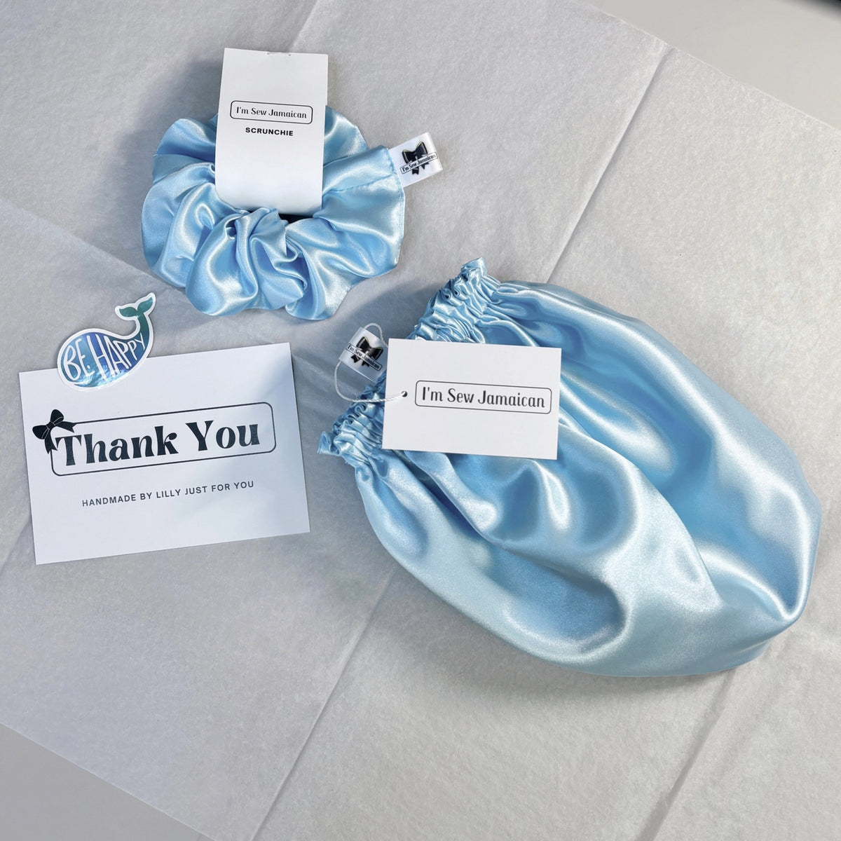 A light blue satin hair scrunchie and matching satin bonnet on a light gray surface, with small white tags reading 'I'm Sew Jamaican' and a 'Thank You' card beside them.