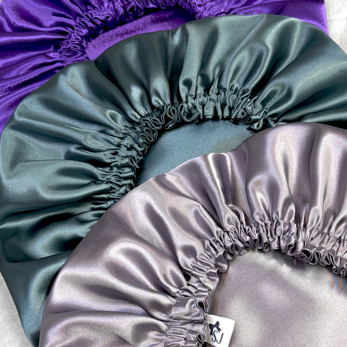 Three satin sleep hats or bonnets in purple, dark teal-green, and light lavender-gray, arranged overlapping on a white background.