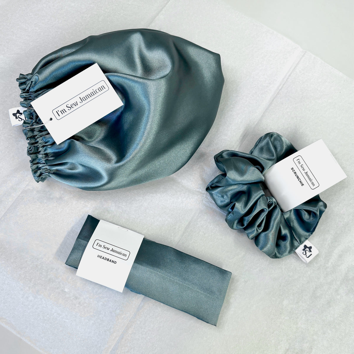Three satin hair accessories in a muted blue-gray color, including a scrunchie, a hair bonnet, and headband.