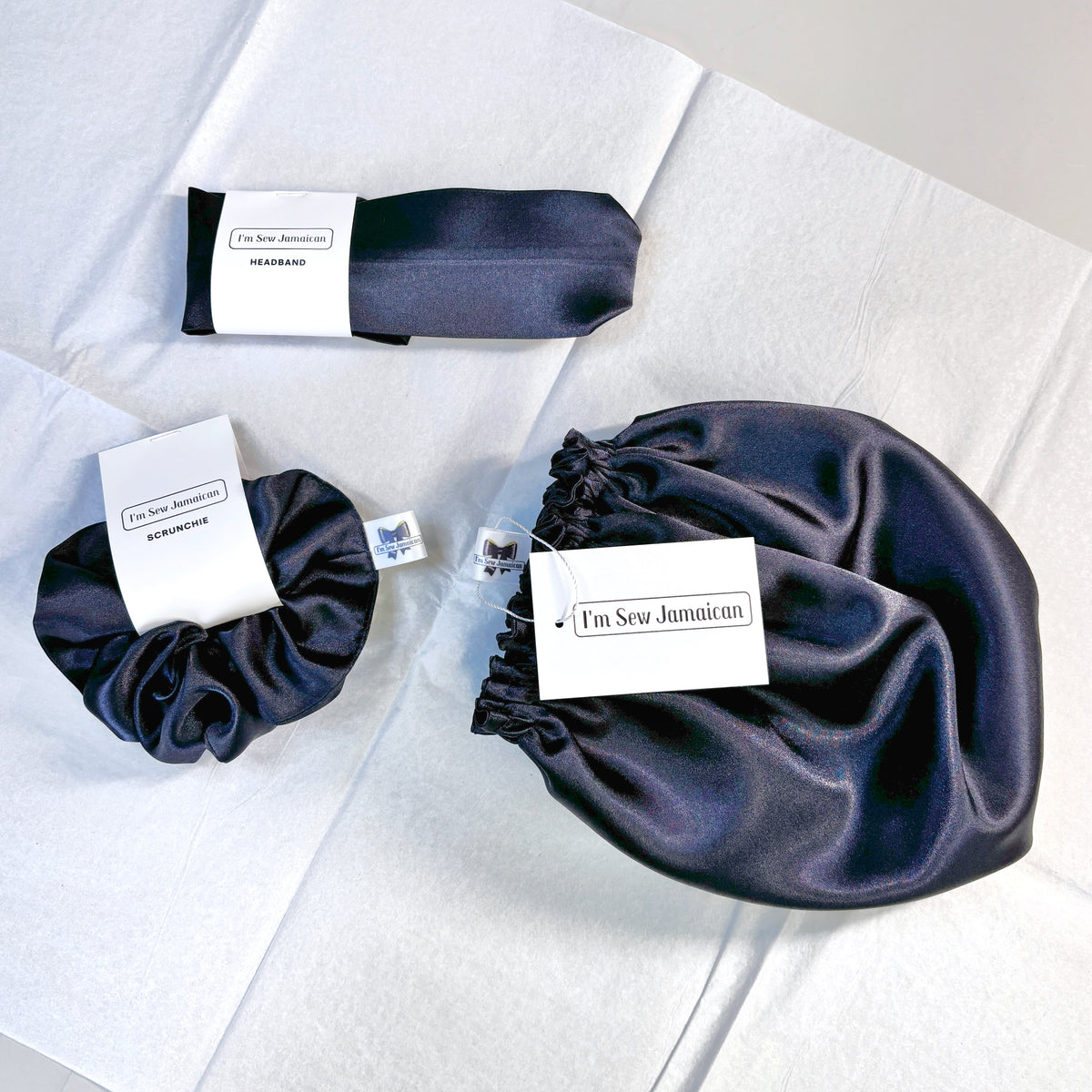 Three dark navy satin hair accessories laid out on white fabric: a scrunchie, a headband, and satin bonnet, each with a white paper tag. 