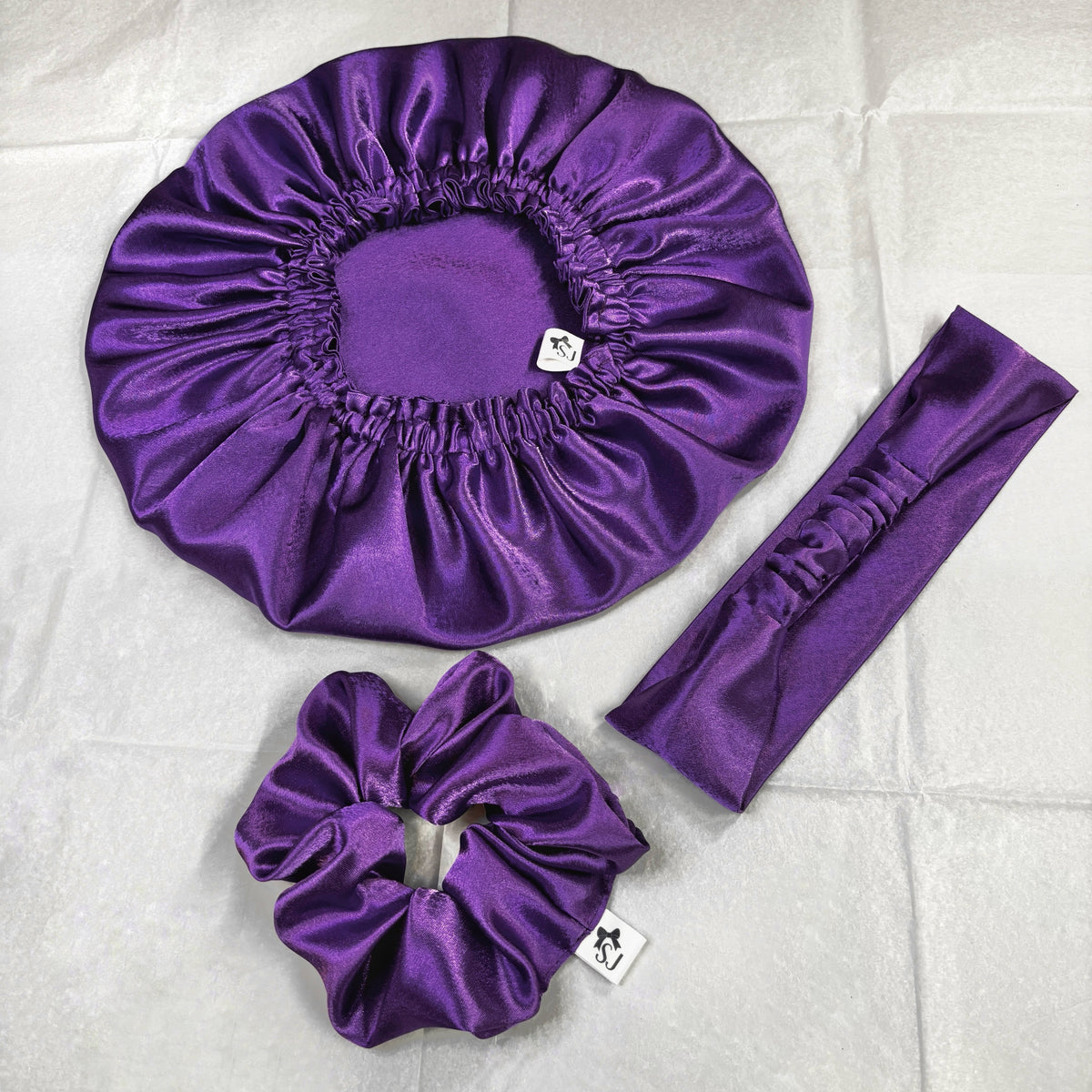 Purple satin hair accessories set including a large bonnet, scrunchie, and headband, laid out on a white fabric background.