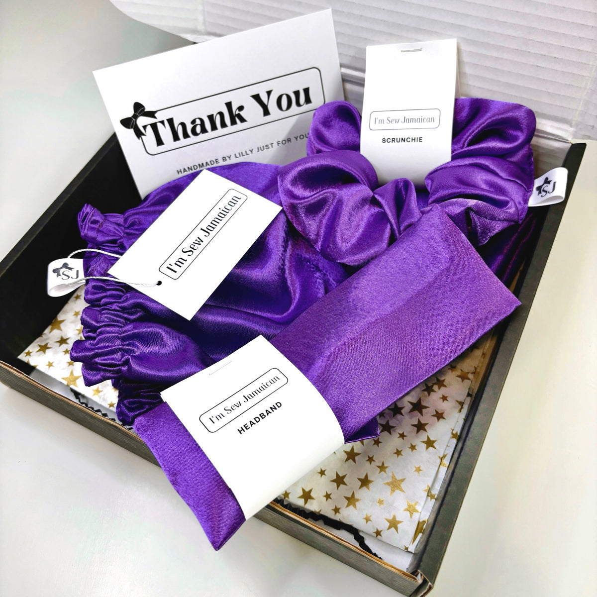 A gift box containing a purple satin scrunchie, satin bonnet and matching headband with white tags displayed on a white background.