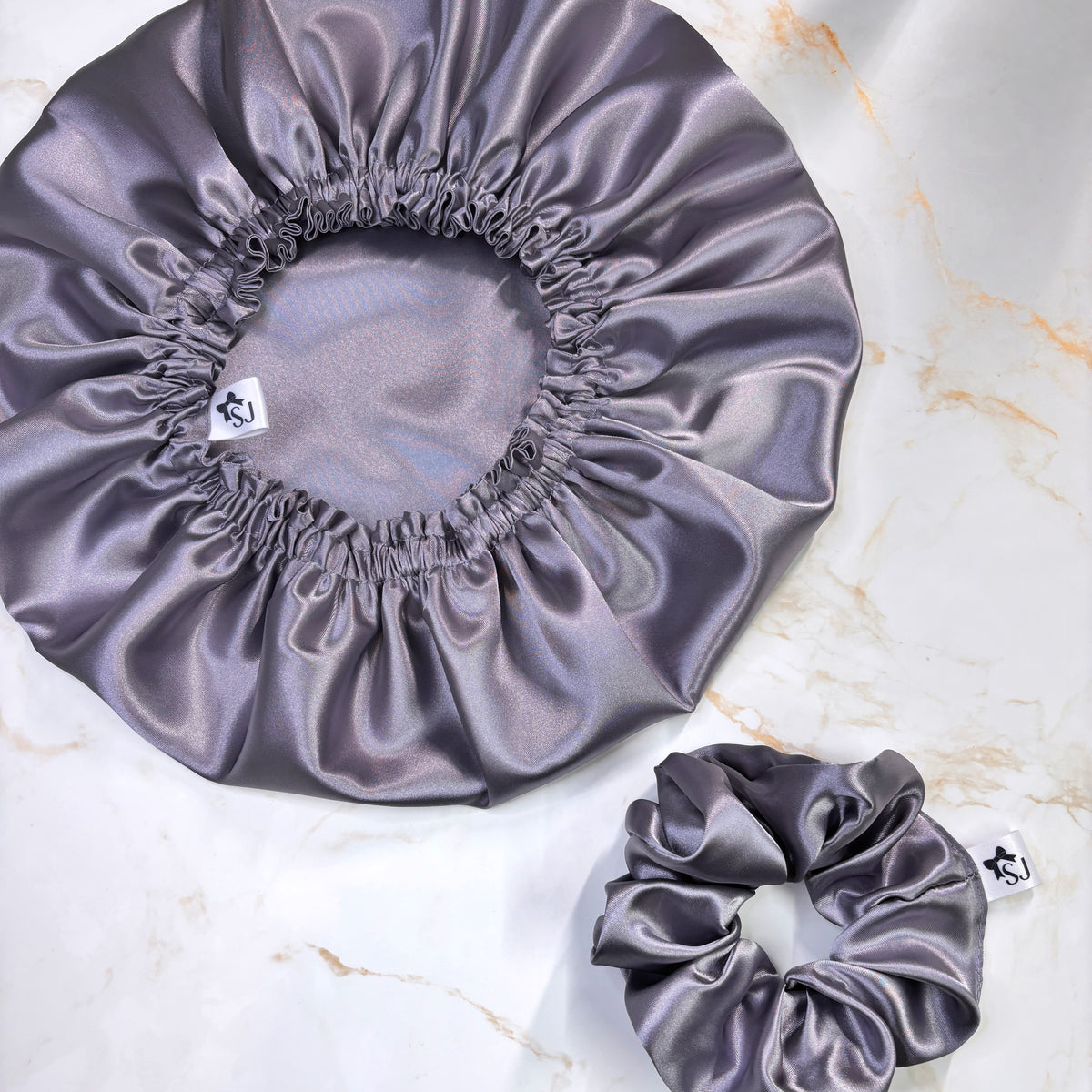 A satin sleep cap and matching satin scrunchie in a muted lavender-gray color, displayed on a white marble background. Each item has a small white brand tag with a black logo.