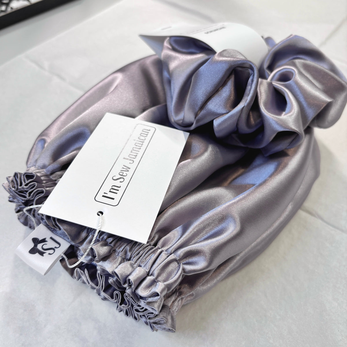 A lavender-purple satin hair scrunchie with an elasticized gathered edge, shown folded on a light fabric background with a white paper tag attached.