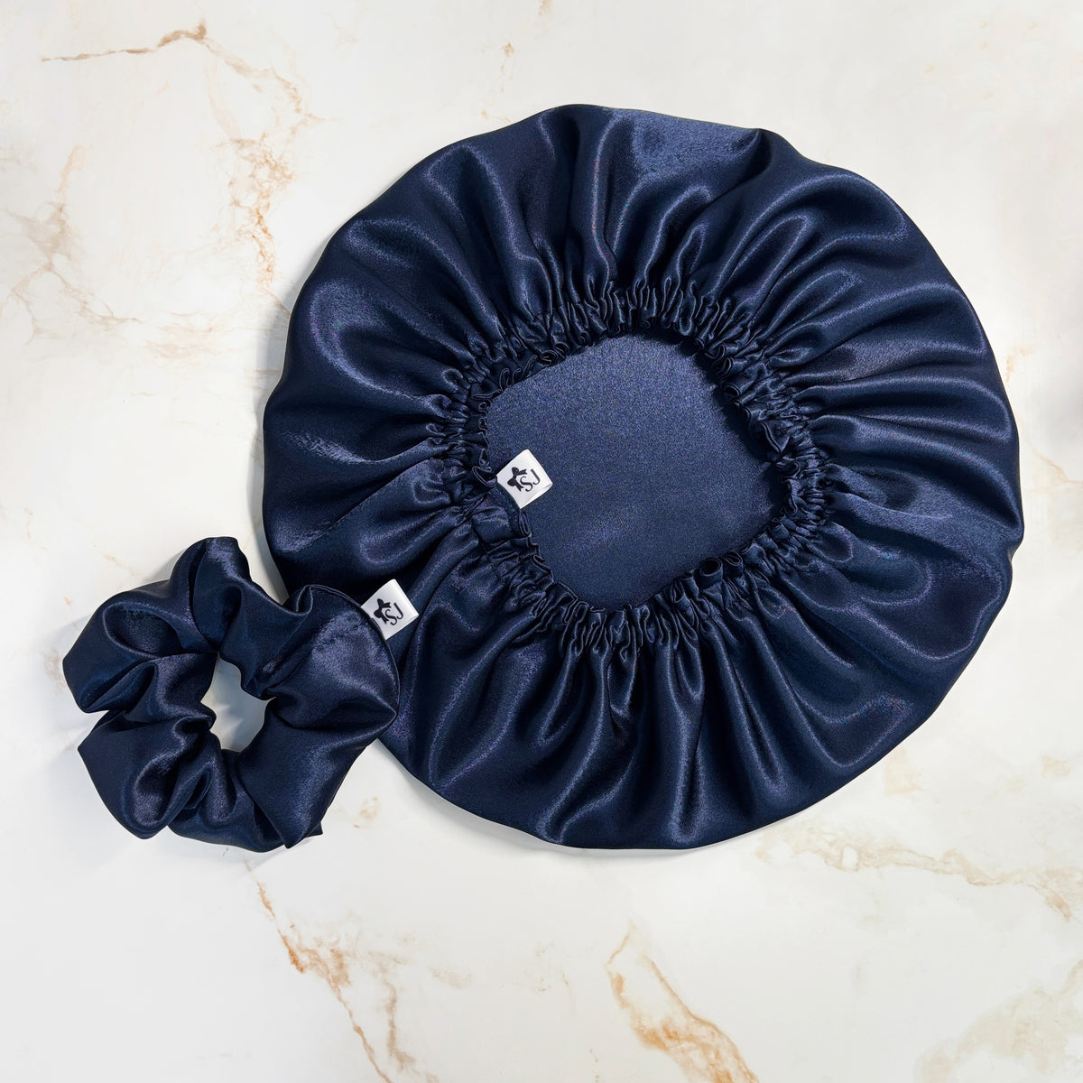 A navy blue satin bonnet and matching scrunchie laid flat on a light beige marble surface. Each bonnet has a small white square tag with a black monogram logo.