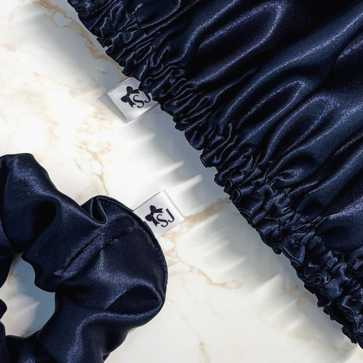 A close-up of a navy blue satin scrunchie and matching elasticized fabric piece with small white tags featuring a black monogram logo, arranged on a light marble surface.