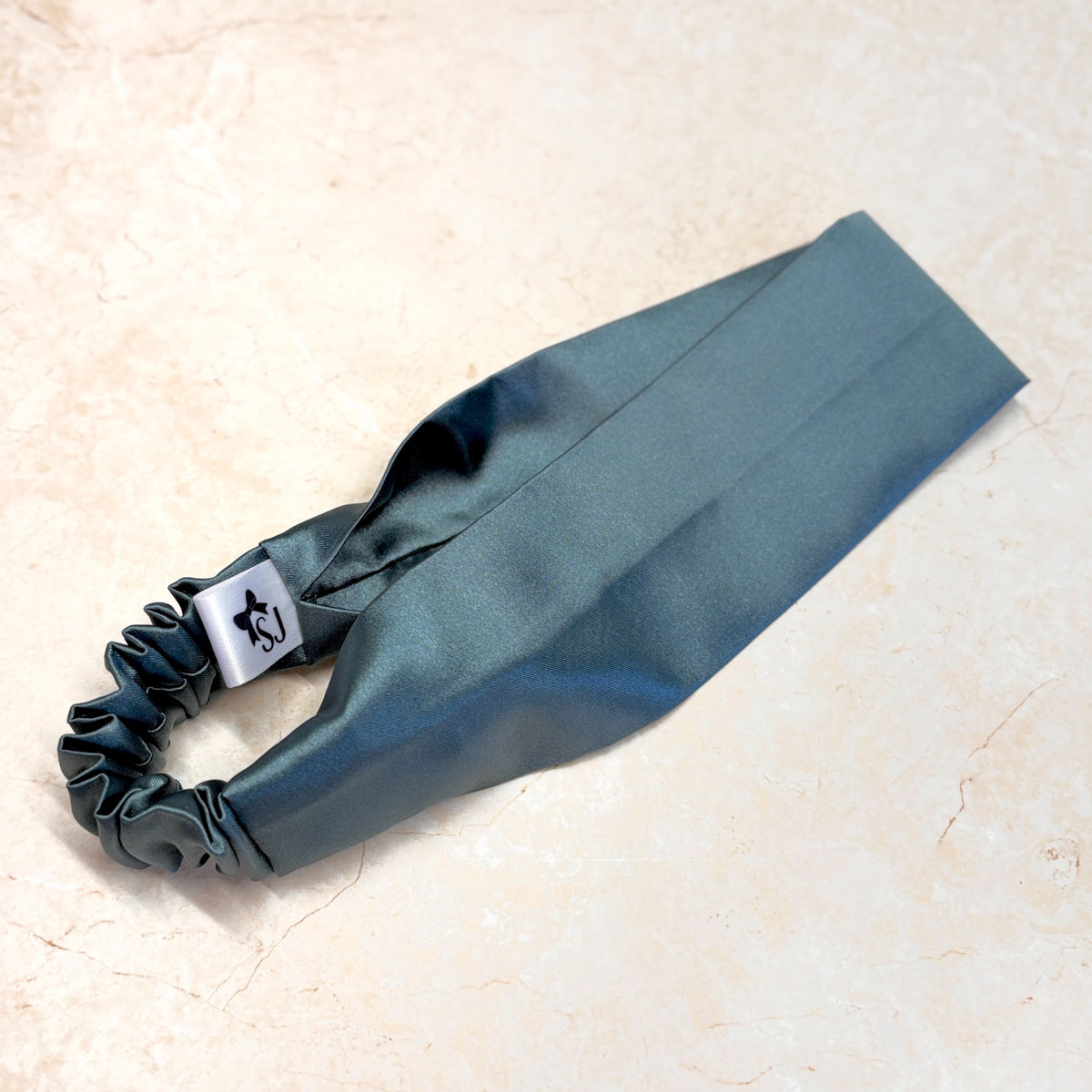 A dark teal satin headband white brand tag, displayed on a light beige marble surface.