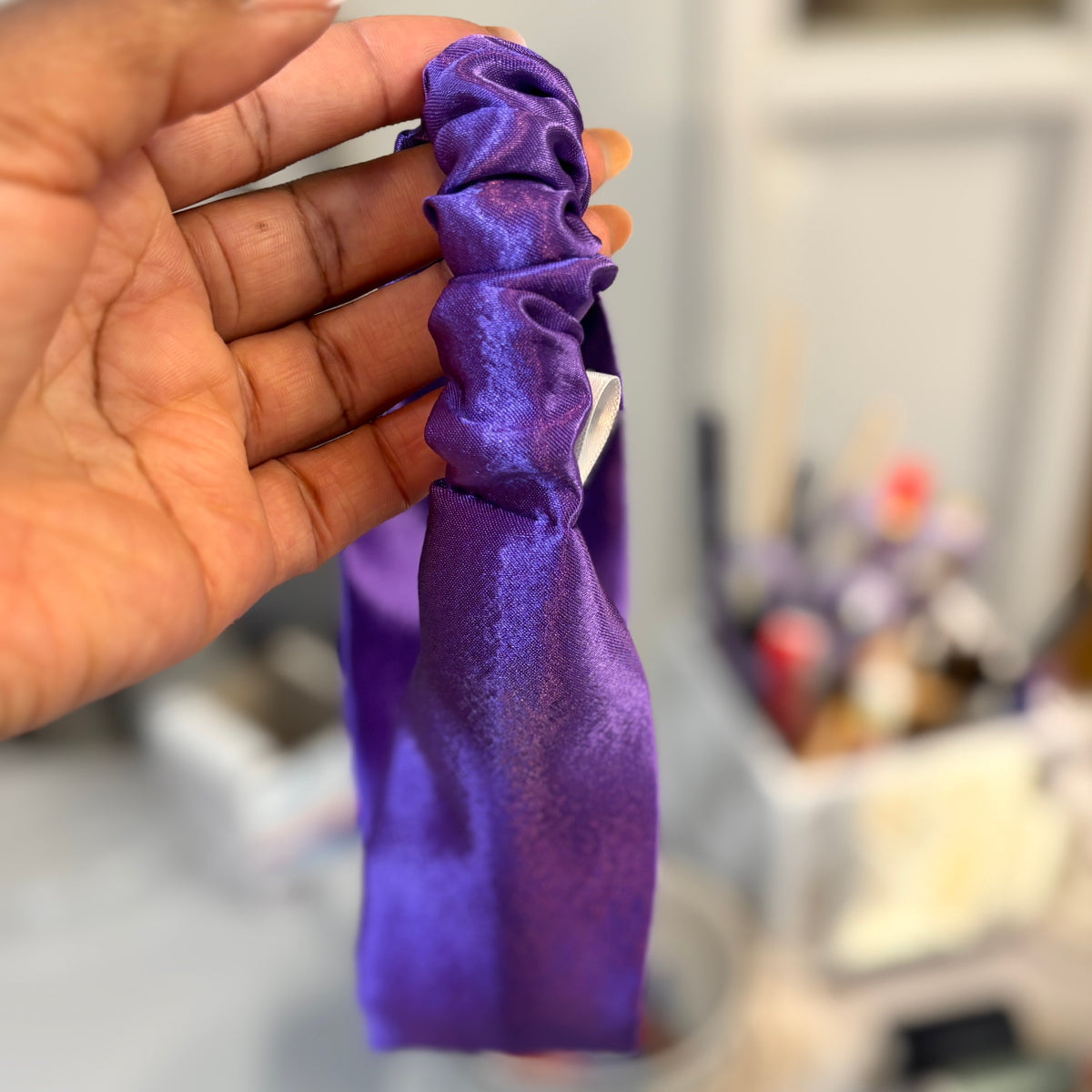 A hand holding a shiny purple satin scrunchie against a blurred indoor background.