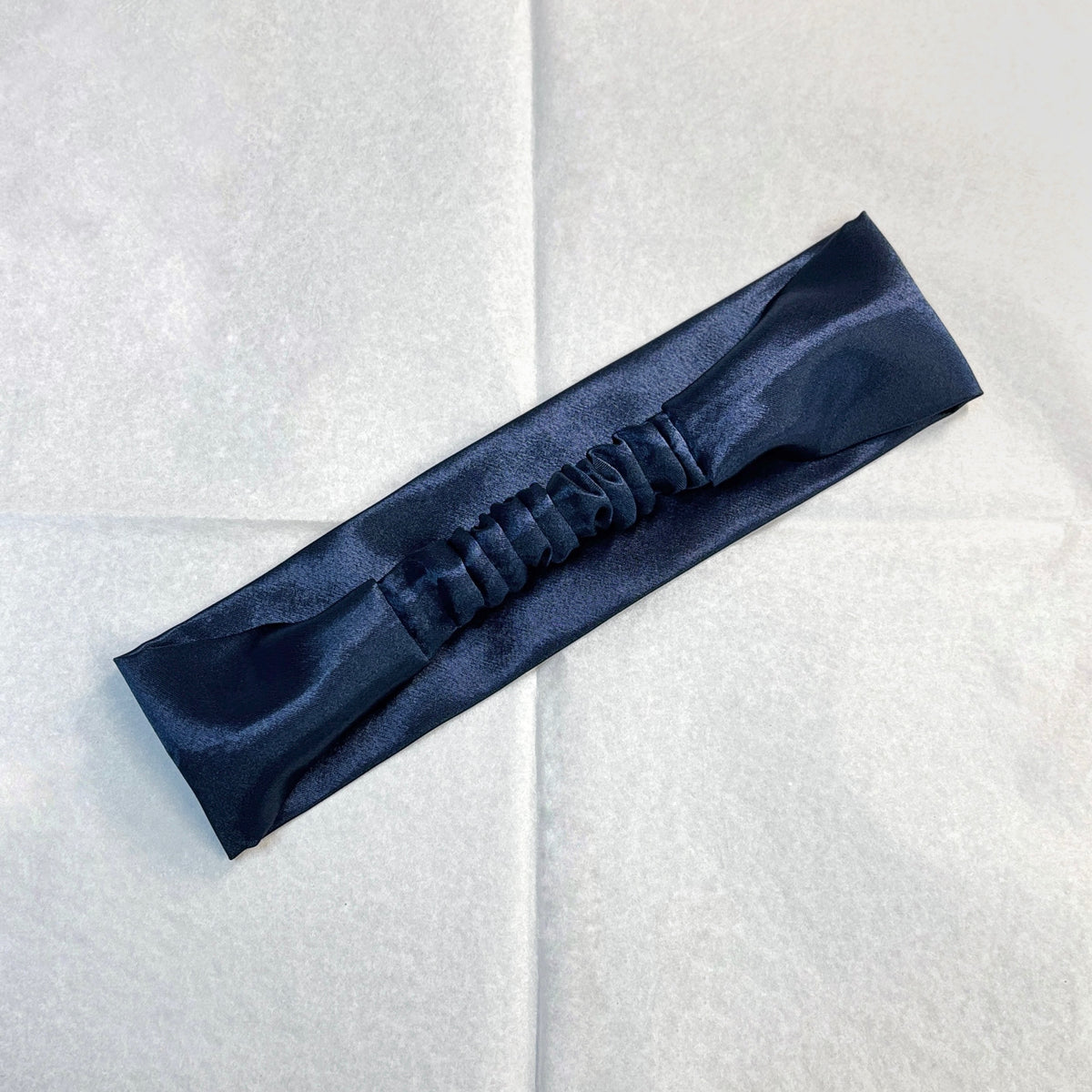 A dark navy blue satin headband with an elasticized center, laid diagonally on a light gray textured background.