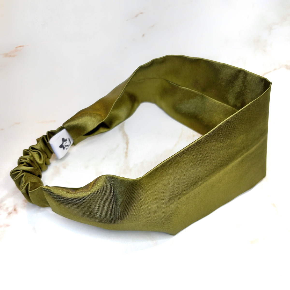 An olive green satin headband with a gathered elastic end and a small white brand tag, displayed on a white marble surface.