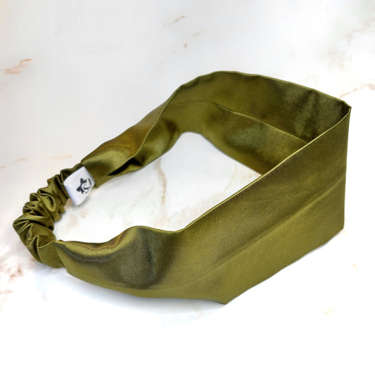 An olive green satin headband with a gathered elastic end and a small white brand tag, displayed on a white marble surface.