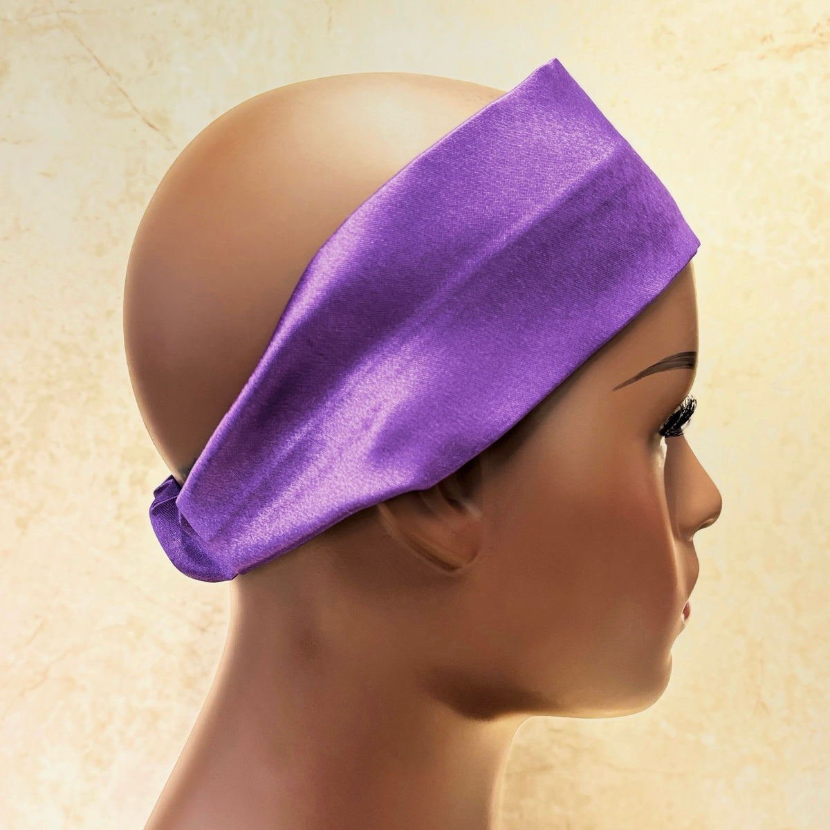 A purple satin headband displayed on a mannequin head in profile against a textured beige background.