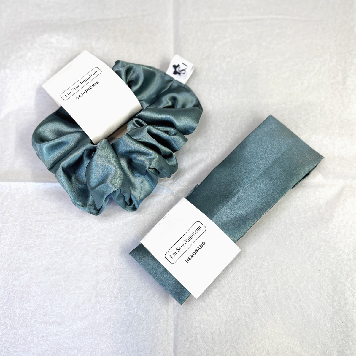 A satin scrunchie and matching headband set in a muted teal-green color, each with a white paper tag, displayed on a textured white fabric background.