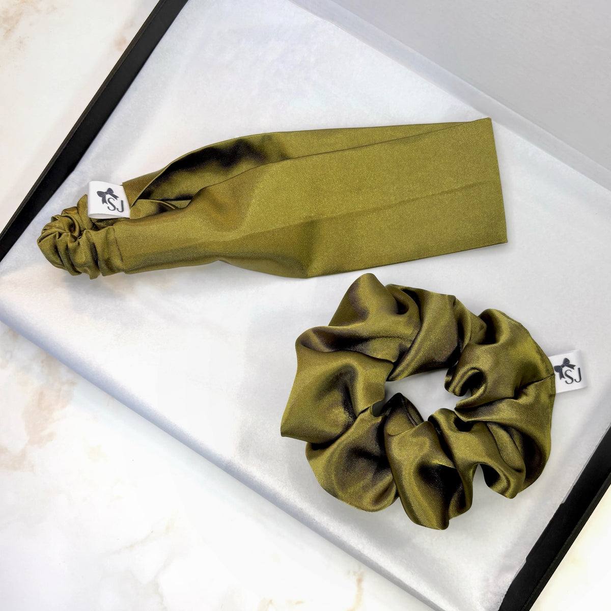 An olive green satin hair accessory set featuring a scrunchie and a matching flat headband both displayed on a white textured surface inside a black box.
