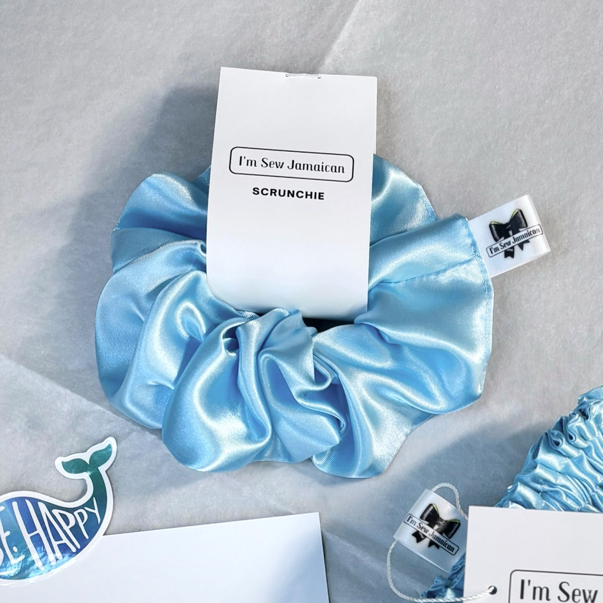 A light blue satin scrunchie with a white paper tag that reads 'I'm Sew Jamaican SCRUNCHIE', displayed on a light gray fabric background with other decorative tags and partial items around it.