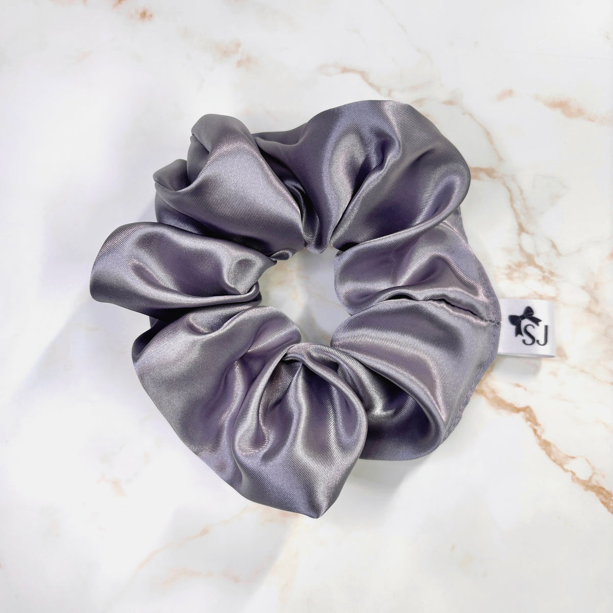 A satin hair scrunchie in a dusty lavender-gray color, laid flat on a white marble surface. A small white brand tag with the letters 'ISJ' is visible on the right side of the scrunchie.