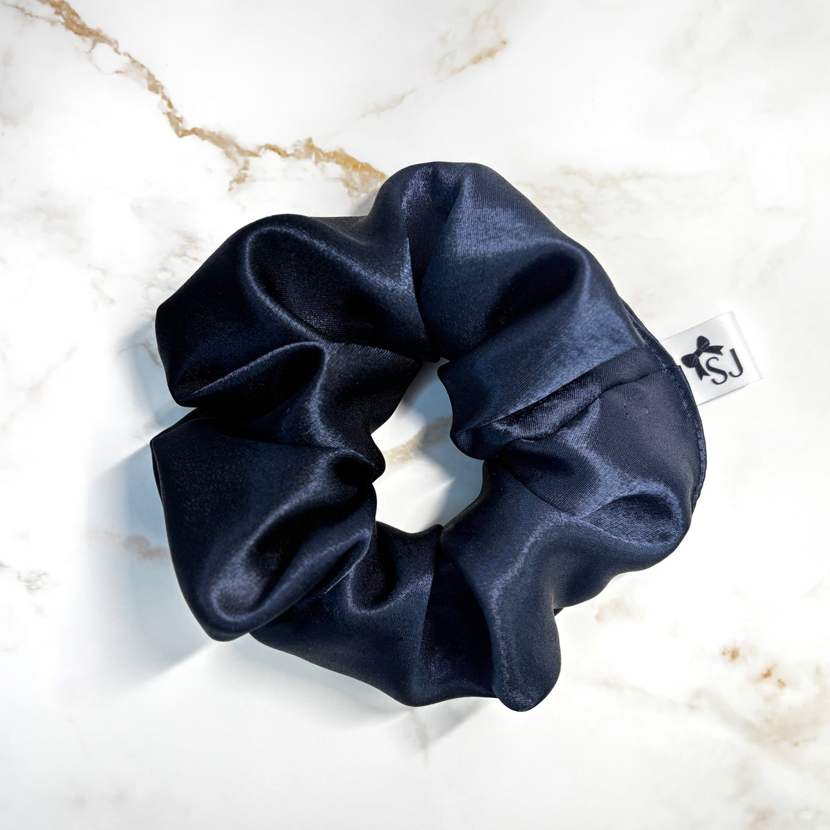 A dark navy blue satin hair scrunchie with a small white brand tag featuring a black star logo, placed on a white marble surface with gold veining.