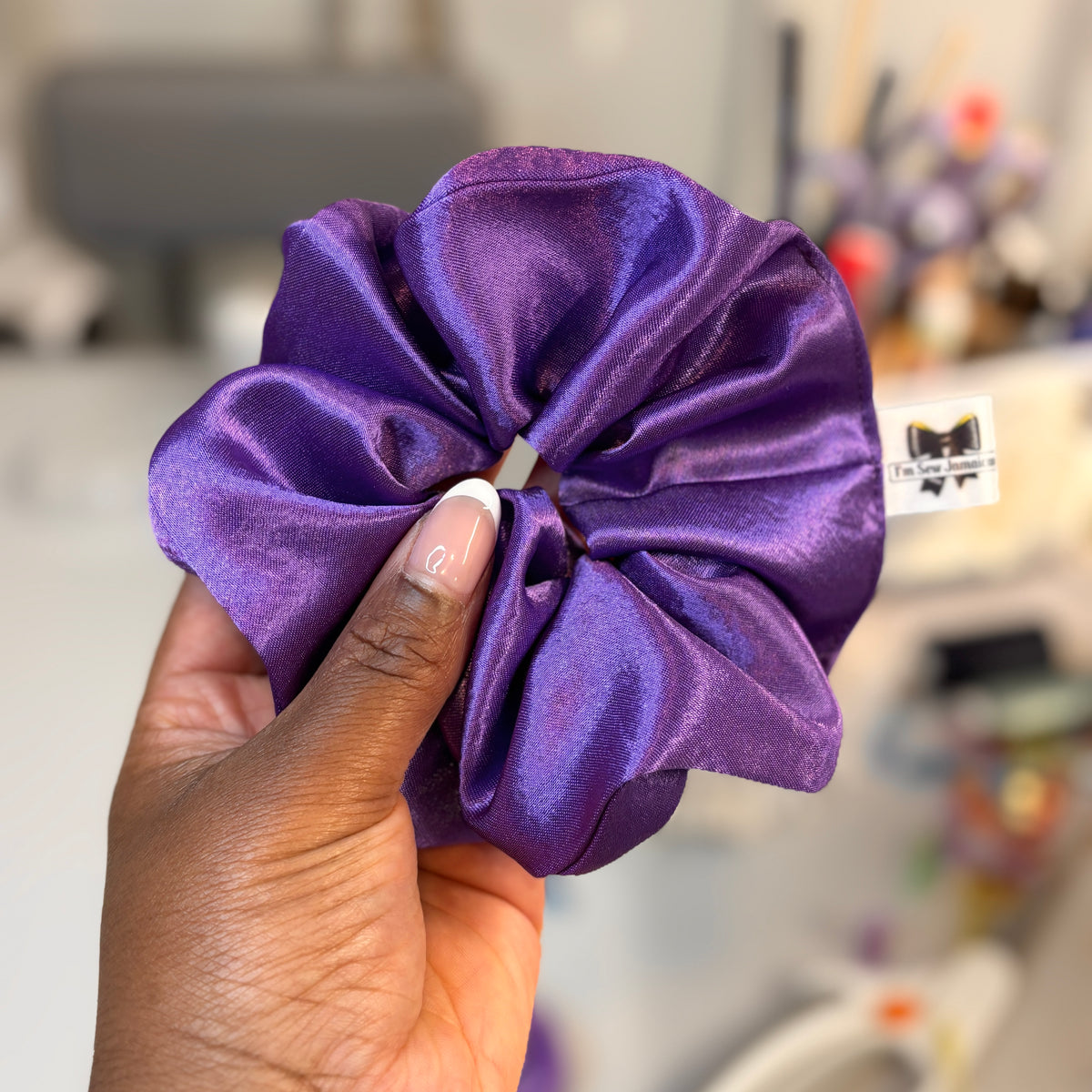 A hand holding a large purple satin hair scrunchie with a small white brand tag attached, set against a blurred indoor background.
